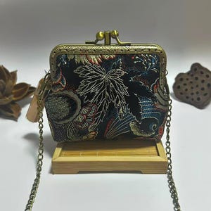 May include: A small, black clutch bag with a decorative floral pattern in red, white, and blue. The purse has a gold-coloured metal frame and chain strap. The clutch is displayed on a small wooden stand.