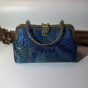 Może przedstawiać: Niebieska kopertówka z ozdobną metalową ramą i zapięciem. Torebka ma wirujący wzór w odcieniach niebieskiego, złotego i srebrnego. Uchwyt ma pasujący złoty kolor. Torebka jest w stylu vintage.