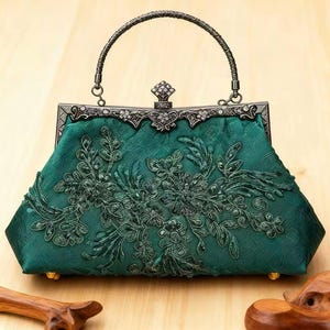May include: Emerald green handbag with intricate floral embroidery and a decorative metal clasp. The bag features a textured fabric and a curved handle. The metal frame is embellished with small, sparkling accents. A stylish accessory for special occasions.