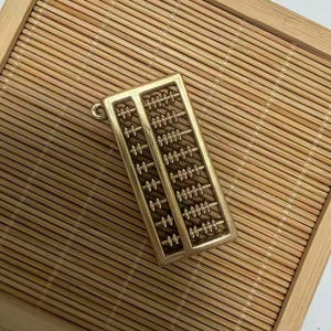 May include: A gold-colored abacus, a traditional calculating tool, rests on a bamboo tray. The abacus has multiple rows of beads, and a small loop is attached to the top. The bamboo tray is rectangular and has a natural wood finish.
