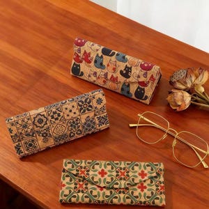 May include: Three cork eyeglass cases with different patterns: one with cat illustrations, one with geometric tiles, and one with a red and green design. A pair of gold-rimmed glasses and dried flowers are also on the wooden surface.