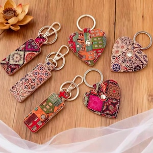 May include: A collection of keychains with colorful, patterned designs. Some are rectangular, others heart-shaped. Each keychain has a silver-colored ring and clasp. The patterns include geometric shapes and house motifs, in shades of red, orange, and purple.