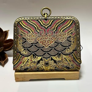 Peut inclure: Pochette carrée ornée d'un cadre en métal doré et d'un fermoir décoratif. Le tissu présente un motif détaillé en noir, or et rouge. La pochette est présentée sur un petit support en bois.