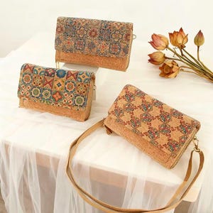 May include: Three cork wallets with colorful geometric patterns. The wallets have a natural cork base and a patterned flap with a zipper closure. One wallet has a long strap. The wallets are displayed on a white surface.