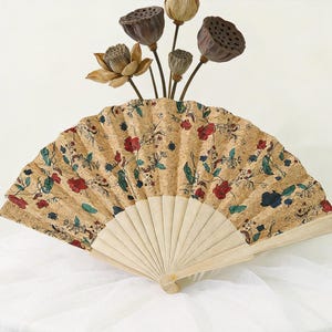 K&ouml;nnte beinhalten: Dekorativer Handf&auml;cher mit korkartiger Oberfl&auml;che, verziert mit einem Blumenmuster in Rot, Blau und Gr&uuml;n. Der F&auml;cher ist ge&ouml;ffnet und zeigt einen hellen Holzrahmen. Getrocknete Lotus-Kapseln und -Bl&uuml;ten sind dar&uuml;ber angeordnet.