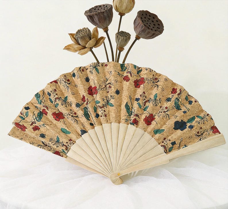 K&ouml;nnte beinhalten: Dekorativer Handf&auml;cher mit korkartiger Oberfl&auml;che, verziert mit einem Blumenmuster in Rot, Blau und Gr&uuml;n. Der F&auml;cher ist ge&ouml;ffnet und zeigt einen hellen Holzrahmen. Getrocknete Lotus-Kapseln und -Bl&uuml;ten sind dar&uuml;ber angeordnet.