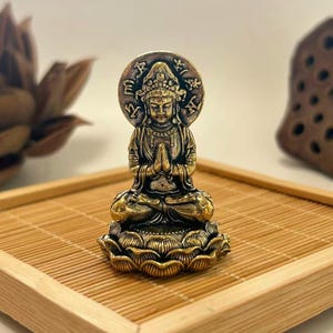 Può includere: Piccola e dettagliata statua di Buddha in ottone in posizione meditativa. La statua è su una base a forma di fiore di loto e ha un'aureola. Si trova su un vassoio di bambù. Simbolo di pace e illuminazione.