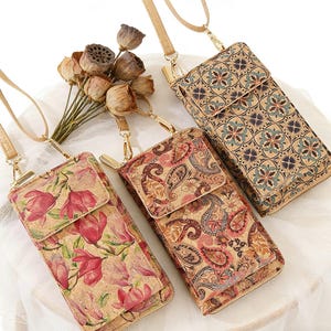 May include: Three cork crossbody phone bags with different floral and paisley patterns. The bags have a flap closure, a zipper, and a long, adjustable strap. The bags are beige with red, pink, and blue accents. A bouquet of dried flowers is in the background.