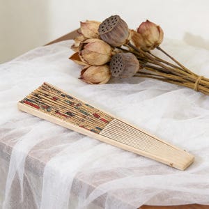K&ouml;nnte beinhalten: Ein h&ouml;lzerner Faltf&auml;cher mit einem dekorativen Druckdesign in Rot, Blau und Gr&uuml;n. Der F&auml;cher ist ge&ouml;ffnet und liegt auf einem wei&szlig;en Tuch. Getrocknete Lotus-Schoten und -Stiele befinden sich im Hintergrund.