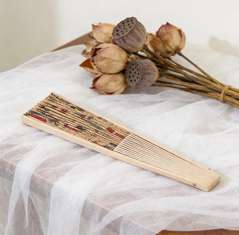 K&ouml;nnte beinhalten: Ein h&ouml;lzerner Faltf&auml;cher mit einem dekorativen Druckdesign in Rot, Blau und Gr&uuml;n. Der F&auml;cher ist ge&ouml;ffnet und liegt auf einem wei&szlig;en Tuch. Getrocknete Lotus-Schoten und -Stiele befinden sich im Hintergrund.