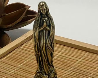 May include: A small, bronze-colored statue of the Virgin Mary in a praying pose. The statue is detailed with flowing robes and a rosary. It stands on a textured base, placed on a bamboo tray.