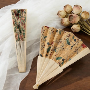 K&ouml;nnte beinhalten: Zwei dekorative Handf&auml;cher. Einer ist geschlossen und zeigt ein farbenfrohes Blumenmuster am Rand. Der andere ist ge&ouml;ffnet und zeigt einen beigen Hintergrund mit einem floralen Design in Rot, Gr&uuml;n und Blau. Beide F&auml;cher haben helle Holzgriffe.