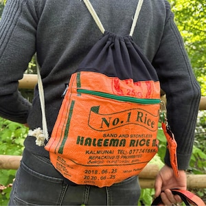Könnte beinhalten: Orange-schwarzer Rucksack mit Kordelzug aus einem recycelten Reissack. Er hat einen grünen Reißverschluss und weiße Seilriemen. Auf der Vorderseite steht der Text "No.1 Rice" und weitere Details.