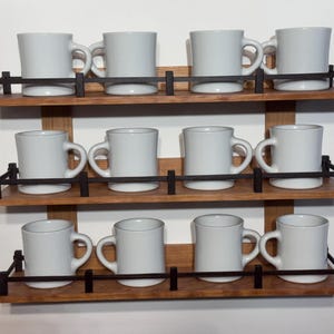 May include: A wooden mug rack with three shelves, each holding four white ceramic mugs. The shelves are made of light brown wood and have black metal railings. The mugs are evenly spaced and have curved handles.