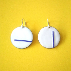 Puede incluir: Un par de pendientes de cerámica blanca con líneas azules. Los pendientes son redondos y tienen un diseño sencillo.