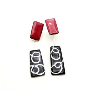 May include: A pair of contemporary earrings. Each earring has a rectangular, glossy, dark red top and a black rectangular bottom with a white, swirled design. The earrings are connected by a silver wire.