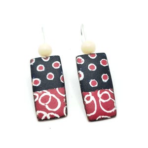 May include: A pair of dangle earrings featuring a black and red geometric pattern. The earrings are made of metal and have a white bead at the top of the dangle.