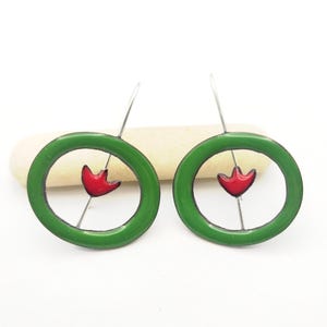 May include: A pair of handmade earrings featuring a green enamel circle with a red tulip-shaped design in the center. The earrings have a silver hook and are set against a white background.