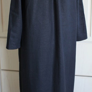 May include: A long, dark blue linen shirt with a button-up collar and long sleeves. The shirt has a single pleat in the back and is hanging on a wooden mannequin.