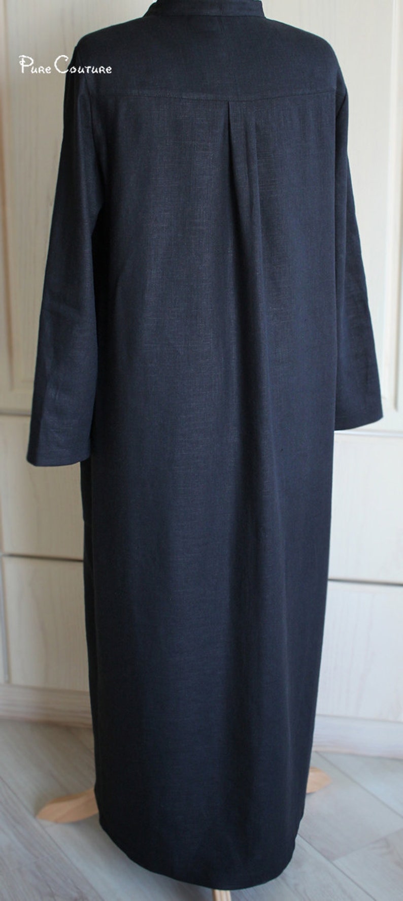 May include: A long, dark blue linen shirt with a button-up collar and long sleeves. The shirt has a single pleat in the back and is hanging on a wooden mannequin.