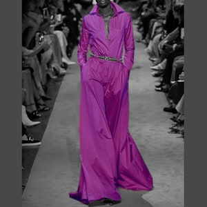 Op de afbeelding: Een levendig paarse jumpsuit met een top in overhemdstijl en broek met wijde pijpen. De outfit heeft lange mouwen, een knoopsluiting aan de voorkant en een bijpassende riem. Het model draagt zwarte schoenen. De achtergrond is een catwalk.