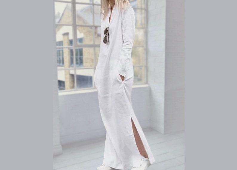 May include: A white long-sleeved maxi dress with a V-neckline and a side slit. The dress has a relaxed fit and is made of a lightweight fabric.