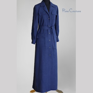 Op de afbeelding: Een lange, donkerblauwe, knoopsluiting jurk met een riem en zakken. De jurk heeft een kraag en lange mouwen. "Pure Couture" staat in witte tekst aan de rechterkant van de afbeelding.