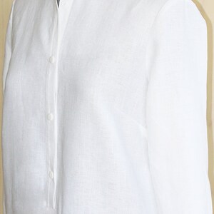 May include: A white linen shirt with a mandarin collar and button closure. The shirt has long sleeves and a relaxed fit.