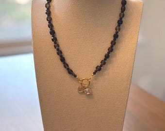 Op de afbeelding: Een ketting met donkerbruine, facetgeslepen kralen. De ketting heeft een goudkleurige sluiting en twee kleine bedels: een zeeschelp en een vierkante bedel. De ketting wordt op een mannequin getoond.