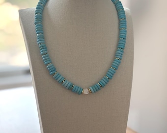 May include: A turquoise beaded necklace with gold-colored spacers and a single, round, white bead. The necklace is displayed on a neutral-colored bust, showcasing its vibrant color and design. The necklace is a stylish accessory.