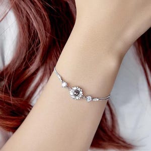 May include: A silver bracelet with a floral centrepiece and clear crystal accents. The bracelet's chain is fine, and it has additional crystals. The bracelet is worn on a wrist. The floral design has a dark stone at its centre.