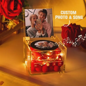 Puede incluir: Una caja de música de acrílico transparente con una foto y el texto "CUSTOM PHOTO & SONG". La caja contiene rosas rojas y luces de hadas. Una foto se muestra en la parte superior y en un disco circular.