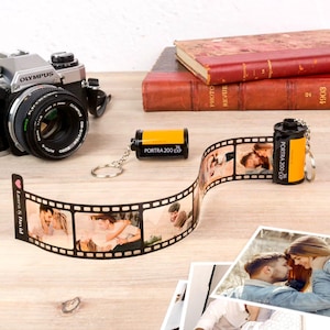 May include: A silver Olympus camera is positioned near a film strip with photos and a film canister keyring. The film strip shows photos of a couple. The keyring has the text "PORTRA 200 EXP".
