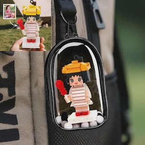 May include: A clear, oval-shaped keychain with a miniature Lego figure inside. The figure is a cartoon character wearing a yellow hat, striped shirt, and red shoes, holding a red microphone. The keychain is attached to a black strap.