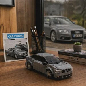 Op de afbeelding: Een grijze Lego Audi-automodel met zwarte accenten staat op een houten oppervlak. Een foto met het label "CAR PHOTO" toont een vergelijkbare auto. Een echte grijze Audi-auto is zichtbaar op de achtergrond.