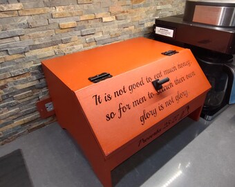 May include: An orange wooden box with a hinged lid and black hardware. The box has a sloped front with the text "It is not good to eat much honey..." and "Proverbs 25:27" in black. The box is on a gray countertop.