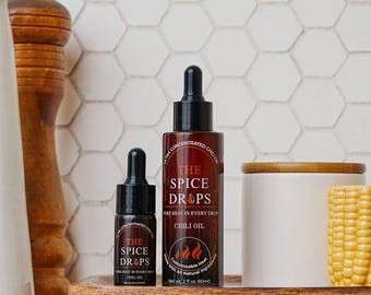 May include: Two bottles of chili oil, one large and one small, with black droppers. The labels read "THE SPICE DROPS" and "PURE HEAT IN EVERY DROP CHILI OIL". The bottles sit on a wooden tray with a white ceramic container and a piece of corn.