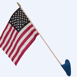 May include: A small United States flag featuring thirteen horizontal red and white stripes and a blue canton with white stars. The flag is attached to a light-coloured wooden pole with a gold-coloured finial and a solid blue plastic base mount.