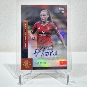 May include: A Topps trading card featuring Ella Toone in a red Manchester United jersey with a blue ink autograph. The card displays the text Manchester United Women, Ella Toone, and the serial number 21/25, all encased in a clear plastic holder.