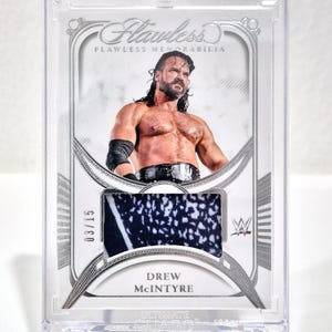 May include: A Drew McIntyre Flawless Memorabilia trading card in a protective case. The card features a photo of Drew McIntyre and a fabric swatch. The card is numbered 03/15.