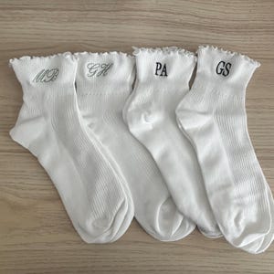 Puede incluir: Cuatro pares de calcetines blancos con un borde superior con volantes. Cada calcetín tiene iniciales bordadas: MB, GH, PA y GS. Los calcetines son de material acanalado y se muestran sobre una superficie de madera clara.