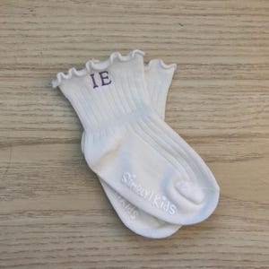 Puede incluir: Un par de calcetines blancos acanalados con un borde superior con volantes. Las letras "IE" están bordadas en morado en el puño. Las palabras "Simply Kids" están impresas en la suela.