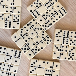 May include: A collection of off-white dominoes with black pips and a small gold-colored metal pin in the center of each tile. The rectangular tiles are arranged in various orientations, displaying different numerical combinations of black dots.