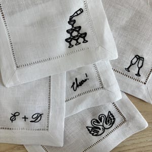 Puede incluir: Servilletas de cóctel de lino blanco con diseños bordados en negro. Los diseños incluyen una torre de champán, dos copas de champán, un par de cisnes y la palabra "Cheers!" en cursiva, junto con las iniciales "E + D".