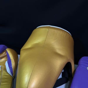 May include: A gold-colored boxing headgear with a white trim and black straps. The headgear is made of a smooth material and is designed to protect the head during boxing or sparring. Other boxing equipment is visible.