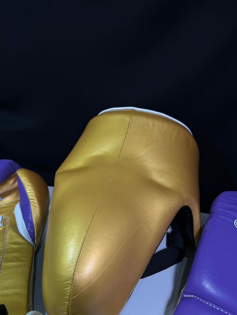 May include: A gold-colored boxing headgear with a white trim and black straps. The headgear is made of a smooth material and is designed to protect the head during boxing or sparring. Other boxing equipment is visible.