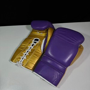 May include: A pair of boxing gloves, featuring a gold and purple color scheme. The gloves are made of leather and have white laces. One glove has "14 oz" printed on it.