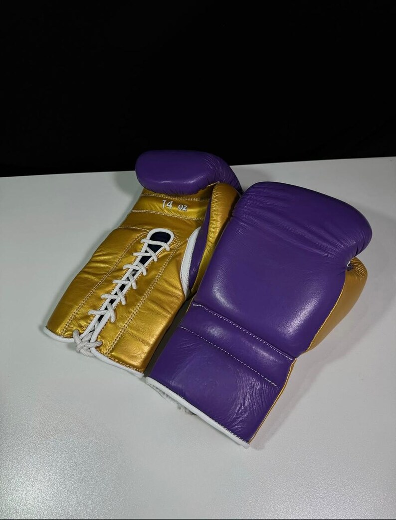 May include: A pair of boxing gloves, featuring a gold and purple color scheme. The gloves are made of leather and have white laces. One glove has "14 oz" printed on it.