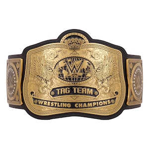 May include: A gold and black wrestling championship belt. The center plate features the WWE logo, a globe, and the words "TAG TEAM" and "WRESTLING CHAMPIONS". The belt has intricate detailing and a crown design.