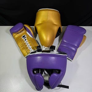 May include: Boxing gear set including a gold headgear, purple headgear, and two boxing gloves. One glove is gold with white laces, and the other is purple. The headgear is gold and purple.